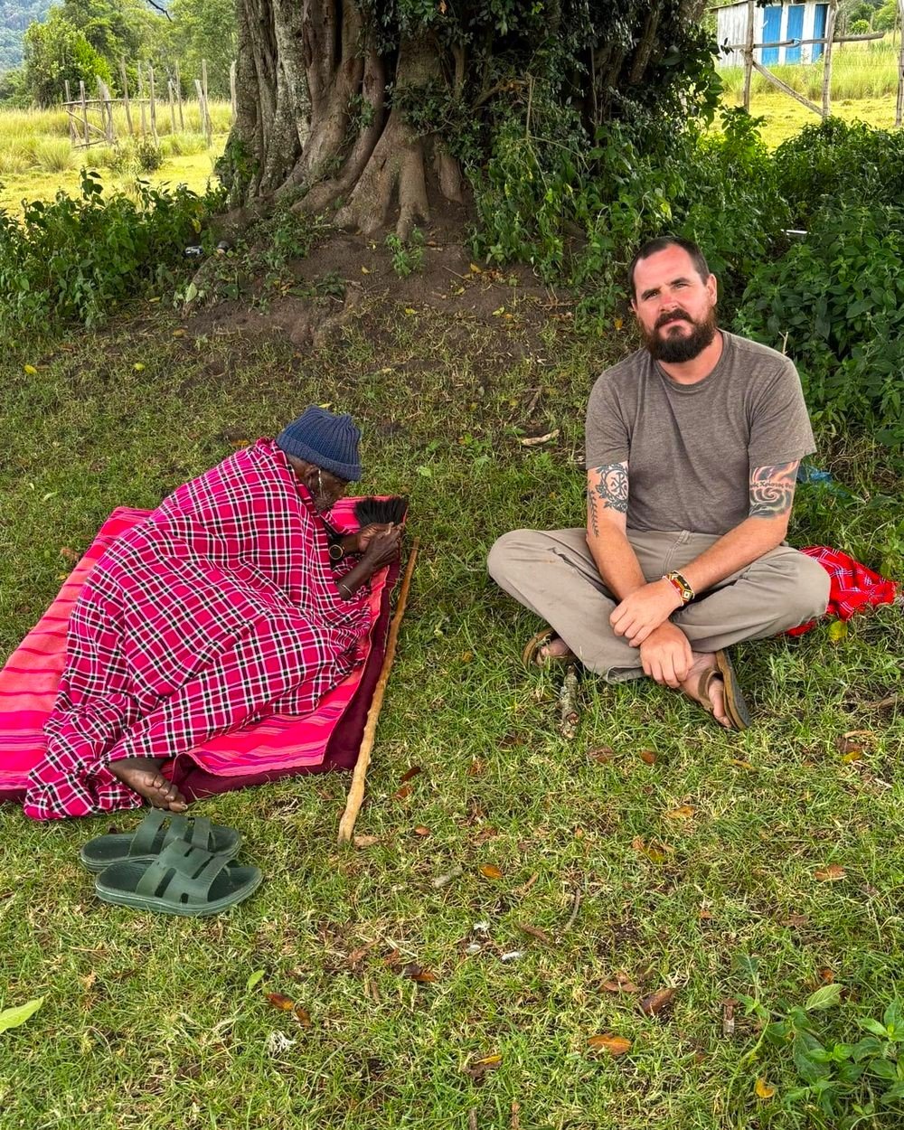 Seth Palmer sitting on the grass beside a Maasai elder wrapped in a red shuka